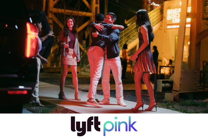 Four young adults greet each other with hugs and smiles at night, near a lit building. A Lyft Pink logo is below the image. 