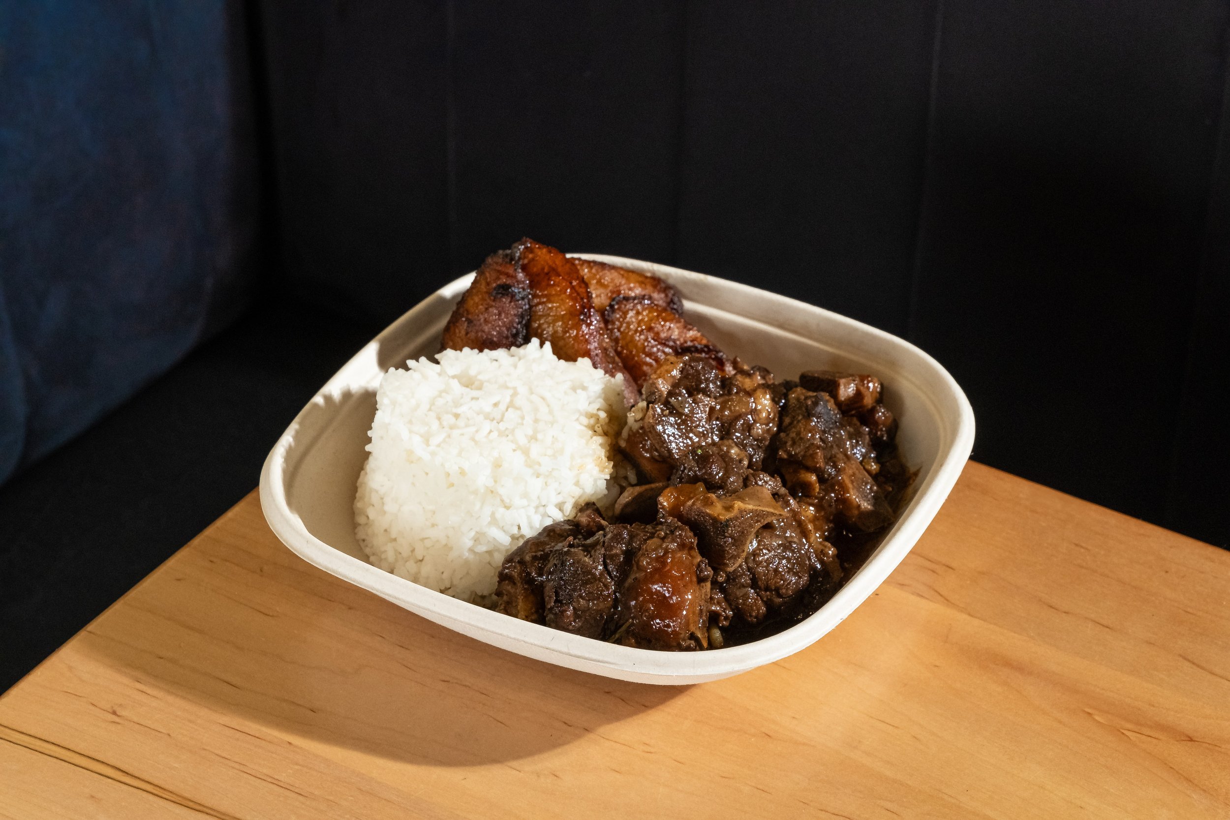 A plate with oxtail, white rice, and fried plantains on a wooden table.