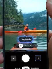 A hand holds a Pixel phone with the camera app open, showing a kayaker on a lake. The zoom function is being used to "Zoom in to 2x."